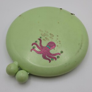 May include: A pale green, circular compact featuring a pink octopus illustration. The octopus has green eyes and is surrounded by small fish and bubbles. Two small, round, pale green knobs are attached.