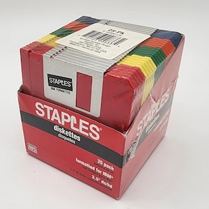 May include: A red box of Staples diskettes, 25-pack, with a variety of colored tabs. The box has the Staples logo and text indicating IBM formatting and 3.5" ds/hd. The box is wrapped in clear plastic.
