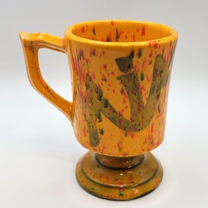 Puede incluir: Una taza de cerámica amarilla con base de pie. La taza tiene un esmalte naranja y rojo moteado con diseños abstractos verdes y marrones.