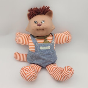 May include: A plush cat doll with a tan vinyl face, brown yarn hair, and blue eyes. The doll wears an orange and white striped long-sleeve shirt, blue denim overalls with a small rectangular label, and a brown collar with a round gold-coloured tag.