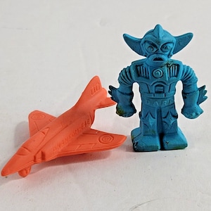 May include: An orange toy airplane with the text "Phantom-FIE" and a blue robot toy. The robot has a detailed design with a stern face and is standing upright. Both toys are made of a rubbery material.