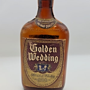 Könnte beinhalten: Eine bernsteinfarbene Glasflasche mit Golden Wedding Blended Whiskey. Das Etikett zeigt den Text "Golden Wedding" in einer verzierten Schriftart, darunter "Blended Whiskey". Die Flasche hat einen Metallverschluss und ein "Pre-War Quality" Siegel.