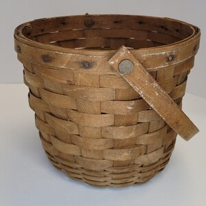 Puede incluir: Una cesta tejida de color marrón con un solo asa de madera. La cesta tiene un aspecto desgastado y rústico y está hecha de materiales naturales.