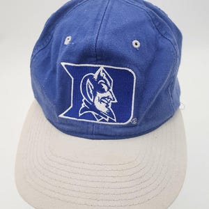 May include: A blue baseball cap with a light gray brim and a top button. The front features an embroidered white Duke University logo with the letter D and a devil mascot profile. The cap has white stitched eyelets on the crown panels.
