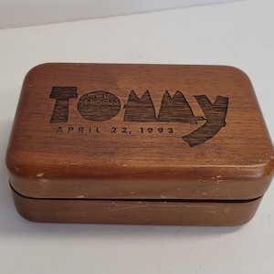 Puede incluir: Una caja de madera con un acabado marrón oscuro. La parte superior de la caja tiene un diseño grabado con las palabras "Tommy" y "The Who's" con la fecha "April 22, 1993".