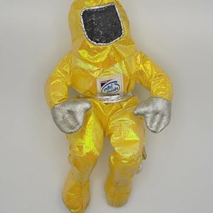 May include: A plush toy in the shape of a yellow hazmat suit with silver gloves and a black face shield. The toy has an "Intel Inside" logo on the chest and a silver belt. The suit is made of a shiny, iridescent material.