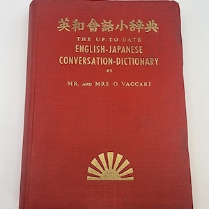 May include: A red, hardcover English-Japanese conversation dictionary. The cover features gold lettering and a gold sunburst design. The text reads "THE UP-TO-DATE ENGLISH-JAPANESE CONVERSATION-DICTIONARY" by Mr. and Mrs. O. Vaccari.