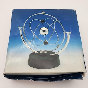 Puede incluir: Una escultura de movimiento cinético con una esfera central plateada rodeada de anillos plateados entrelazados y esferas negras más pequeñas. El dispositivo está montado sobre una base circular negra y se exhibe sobre una caja de cartón azul y blanca.
