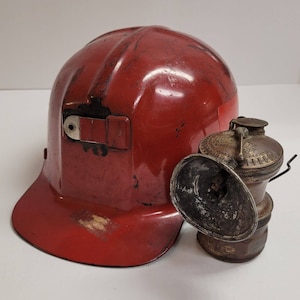 Può includere: Un casco da minatore rosso vintage e una lampada da minatore in ottone. Il casco è di un rosso intenso con segni di usura. La lampada ha un riflettore ed è contrassegnata con testo. Gli oggetti provengono probabilmente dall'industria mineraria.