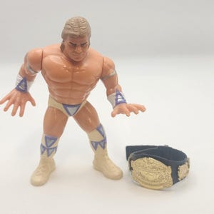 May include: A plastic action figure of a muscular wrestler with blonde hair, wearing cream-coloured trunks, wristbands, and boots with purple triangular accents. Beside the figure is a black championship belt with a gold-coloured metal plate.
