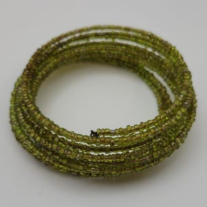 May include: A coiled bracelet made of small, olive-green beads. The beads are strung together to form multiple strands that wrap around each other. The bracelet is set against a plain white background.