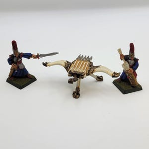 May include: Miniature figurines of soldiers in blue and red uniforms, with swords and a crossbow-like weapon. The figures are on green bases, and the crossbow is off-white and brown.