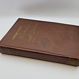 May include: A brown hardcover book titled "Chemistry in Health and Disease" with gold lettering. The book is lying on a white surface. The book's cover is slightly worn, suggesting it is vintage.