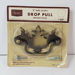 May include: An antique finish drop pull cabinet hardware from Sears. The hardware is 3 inches in hole center and includes two screws. The drop pull has an oval backplate with a decorative leaf design and a brass finish.