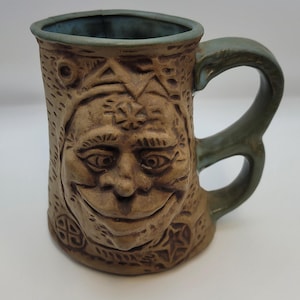 Puede incluir: Una taza de cerámica hecha a mano con una cara sonriente tallada en el exterior de color marrón claro. El asa y el borde son de un verde-azul apagado. La taza presenta tallas en relieve detalladas de formas geométricas y una cara sonriente.