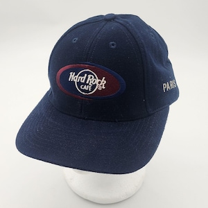 May include: A navy blue baseball cap featuring an embroidered Hard Rock Cafe logo in white and maroon on the front panel. The word PARIS is embroidered in white on the side of the cap. The hat has a structured crown and a flat, stiff brim.