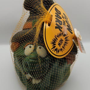 May include: A green ceramic garden gnome with a black hat and white eyes, packaged in a yellow mesh net. The gnome has a tag that reads "Glow in the Dark" and "NEW".