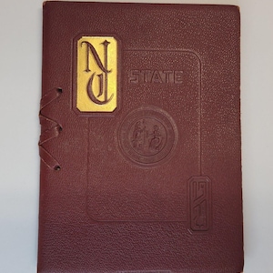 Op de afbeelding: Een in donkerrood leer gebonden boek met gouden letters die "N.C. STATE" lezen. Het boek heeft een gouden zegel met een ontwerp van drie figuren en de tekst "North Carolina State College of Agriculture and Mechanic Arts".
