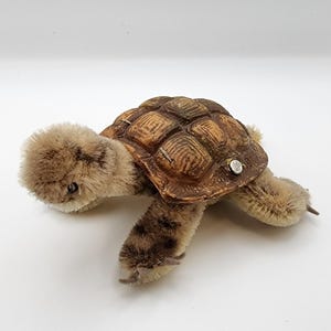 May include: A plush turtle toy with a brown and tan color scheme. The turtle has a textured shell and soft, fuzzy body parts. The toy has a button on its shell and is lying on a white surface.