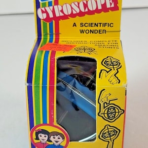 Puede incluir: Una caja de juguetes Gyroscope Steven vintage, amarilla con rayas azules, rojas y verdes. La caja presenta el texto "GYROSCOPE" y "A SCIENTIFIC WONDER". Incluye instrucciones para acrobacias increíbles. A partir de 5 años. Nº 952.