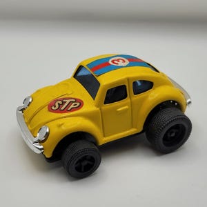 Pode incluir: Um carro de brinquedo amarelo com pneus pretos e o logotipo STP na frente. O teto do carro tem listras azuis e vermelhas com o número 3. O carro tem um para-choque e faróis prateados.