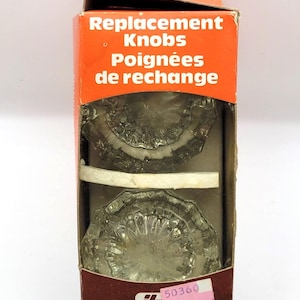 Peut inclure: Une boîte de boutons de remplacement. La boîte orange et marron indique "Replacement Knobs Poignées de rechange". Deux boutons en verre transparent sont visibles à l'intérieur de la boîte. La boîte porte une étiquette de prix avec la marque "UNICAN".