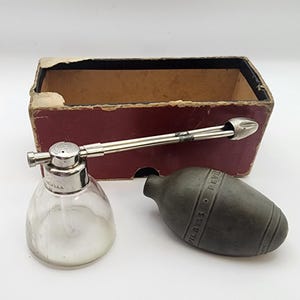May include: A vintage DeVilbiss atomizer with a clear glass base, a silver-toned metal nozzle, and a dark gray rubber bulb. The device is accompanied by a rectangular cardboard box with a maroon exterior and a tan interior lining.
