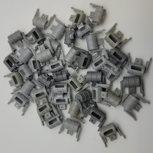 May include: A collection of small, gray plastic model parts, likely for a miniature project. The parts have a mechanical appearance with cylindrical and rectangular shapes, rivets, and other details. The pieces are scattered on a white surface.