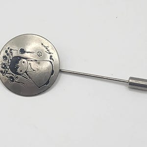 May include: A circular silver-toned lapel pin featuring an etched illustration of a person with dark hair wearing a floral headpiece. The design includes the script text "le gopie" etched into the metal surface. The pin has a long, thin metal stem.