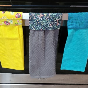 May include: Three kitchen towels hanging on a rack. The towels are yellow with red ladybugs, gray with a floral pattern, and turquoise with a paisley pattern.