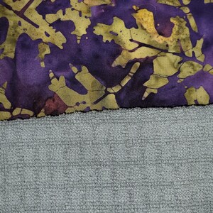 May include: A purple and green fabric with a batik pattern of leaves and branches. The fabric is sewn to a grey textured fabric.