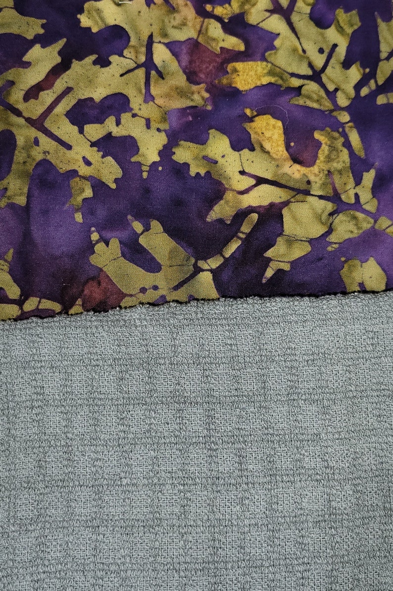 May include: A purple and green fabric with a batik pattern of leaves and branches. The fabric is sewn to a grey textured fabric.