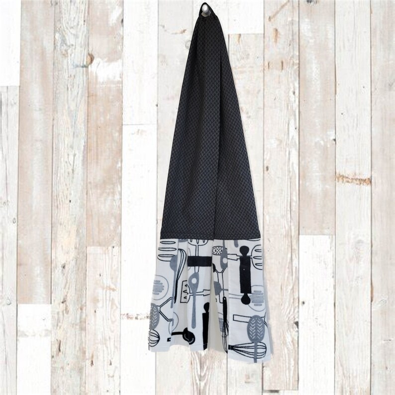 May include: A black and white kitchen towel with a pattern of kitchen utensils. The towel is hanging on a hook. The towel is made of a soft, absorbent material.