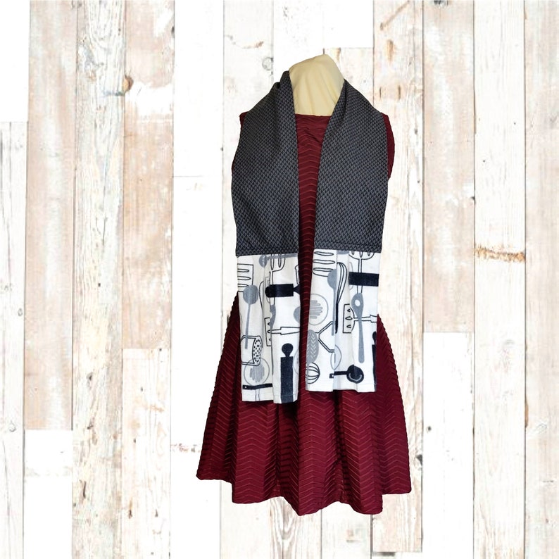 May include: A grey and white kitchen towel with a black and white pattern of kitchen utensils. The towel is hanging on a wooden mannequin wearing a maroon dress.
