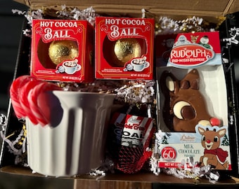 May include: A gift box filled with holiday treats. It includes two boxes of "Hot Cocoa Ball", a white mug with red and white candy canes, a chocolate Rudolph the Red-Nosed Reindeer, and a bag of "Double Crisp COAL".