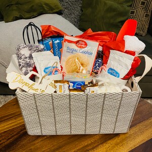 May include: A gift basket filled with baking supplies and treats. Includes a sugar cookie mix, hot chocolate packets, chocolate bars, candy canes, and a whisk. The basket has a gray and white chevron pattern and a wooden tag that says "Donahue 2026".