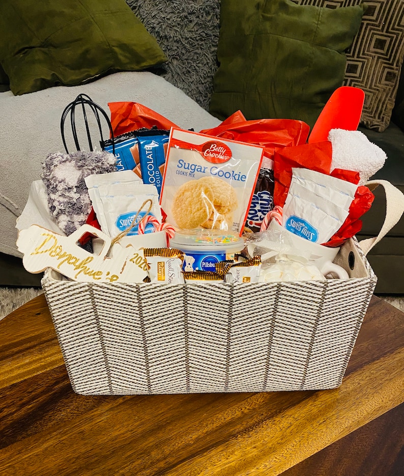May include: A gift basket filled with baking supplies and treats. Includes a sugar cookie mix, hot chocolate packets, chocolate bars, candy canes, and a whisk. The basket has a gray and white chevron pattern and a wooden tag that says "Donahue 2026".