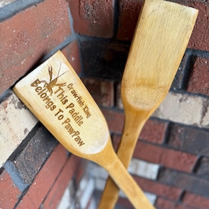 May include: Two wooden paddles with a light brown finish are crossed against a brick wall. One paddle has an engraved crawfish and the text "Crawfish King, This Paddle Belongs To PawPaw."