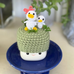 May include: A whimsical ceramic mushroom-shaped planter with a light green crocheted cap, a smiling face, and a blue pedestal base. Two crocheted white ducklings with yellow beaks and feet, and a pink bow, sit atop the cap. Green sprigs add a natural touch.