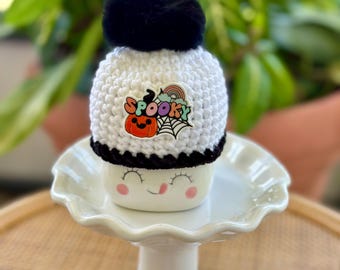 May include: A white crocheted hat with a black pom-pom and a black brim. The hat features a "Spooky" patch with a pumpkin, spiderweb, and rainbow. The hat is displayed on a white ceramic pedestal with a smiling face.