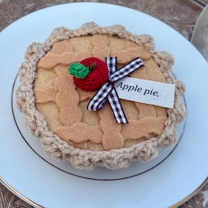May include: A felt apple pie on a white plate. The pie has a lattice crust, a crocheted apple, and a ribbon. The text "Apple pie." is on a small tag. The pie is surrounded by a crocheted border.