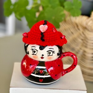 Puede incluir: Una taza de cerámica con forma de mariquita con un sombrero rojo de ganchillo y un adorno de corazón rosa. La taza tiene un cuerpo a rayas blanco y negro y manchas rojas.