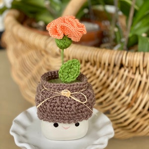 以下が含まれることがあります： 茶色の編み物で覆われた、編み物の花瓶に、桃色の花が咲いています。花瓶は白いケーキスタンドの上に置かれています。