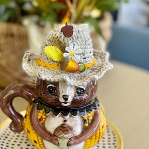 May include: A ceramic squirrel mug with a crocheted hat and fall-themed decorations. The mug is holding a small acorn and is sitting on a black and white polka dot saucer.