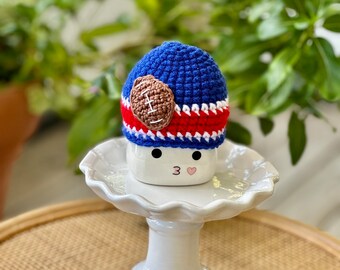 May include: A blue, red, and white crocheted hat with a brown football applique. The hat is placed on top of a white ceramic marshmallow with black eyes and a pink heart. The marshmallow sits on a white, scalloped-edge cake stand.