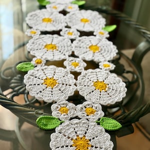 May include: A white crocheted daisy table runner with yellow centers and green leaves. The daisy pattern is repeated throughout the runner, creating a floral design. The runner is placed on a glass table.