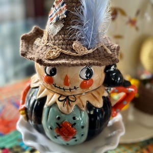May include: A ceramic scarecrow-themed teapot with a brown crocheted hat, a blue feather, and a painted face. The teapot has a black body, a green front, and a white pedestal base. The handle and spout are orange with red polka dots.