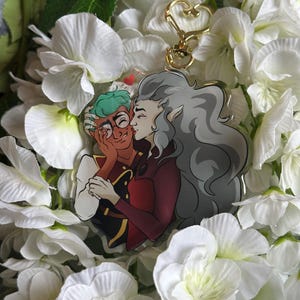 May include: A clear acrylic keychain featuring a cartoon couple kissing. The characters are a green-haired person with glasses and a gray-haired person with elf ears. The keychain is surrounded by white flowers.
