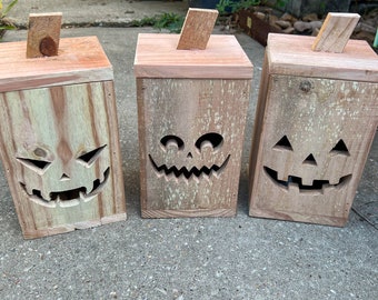 May include: Three wooden jack-o'-lantern boxes with carved faces. The boxes are made of natural wood with a rustic finish. The boxes are arranged in a row on a concrete surface.