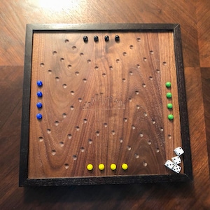 Può includere: Un tabellone da gioco in legno con una cornice nera e una superficie marrone. Ci sono dei buchi nel tabellone e delle biglie blu, verdi, gialle e nere. Ci sono anche tre dadi bianchi sul tabellone.